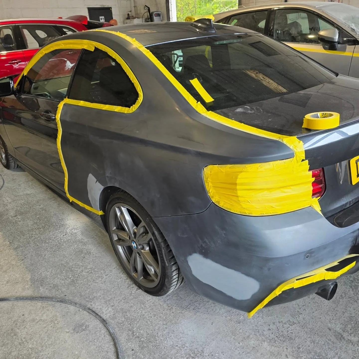 Grey BMW coupé in the workshop masked up with yellow tape around the windows and rear light, with patches of primer on the rear quarter and bumper.