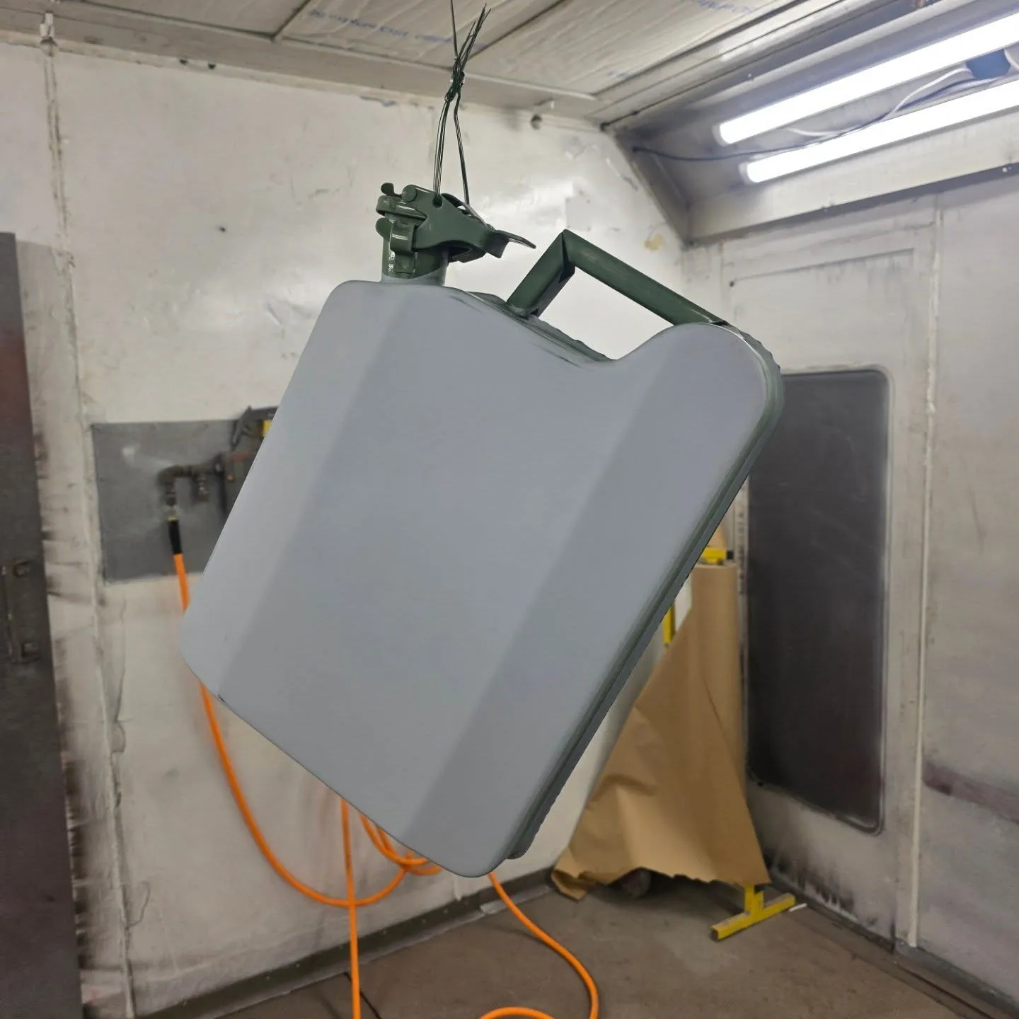 Steel jerry can hanging from a wire in the spray booth, its body freshly painted in pale grey with the green spout and handles left original.