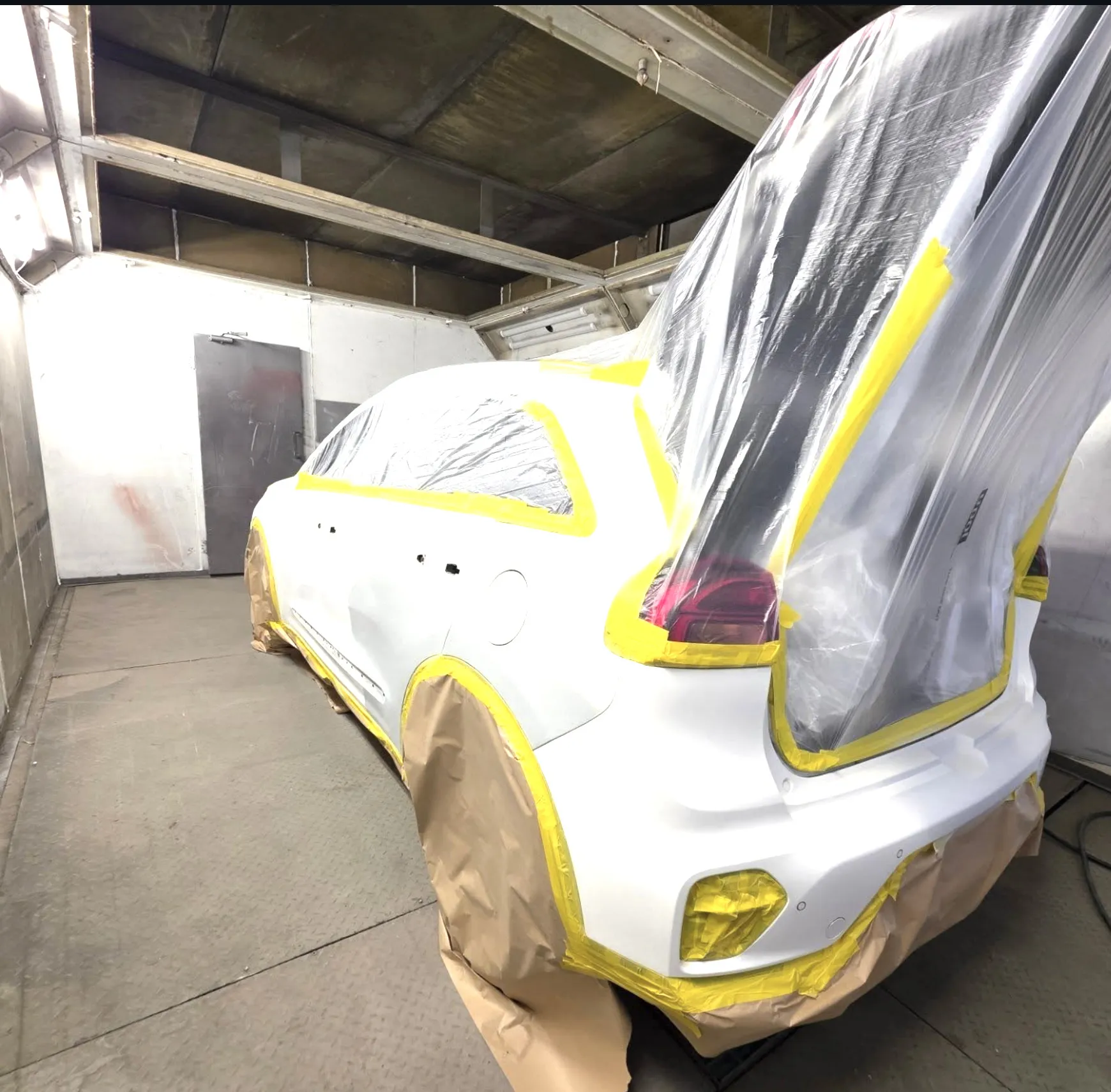 Pearl white Kia masked up with yellow tape and paper, standing in the spray booth with the boot open.