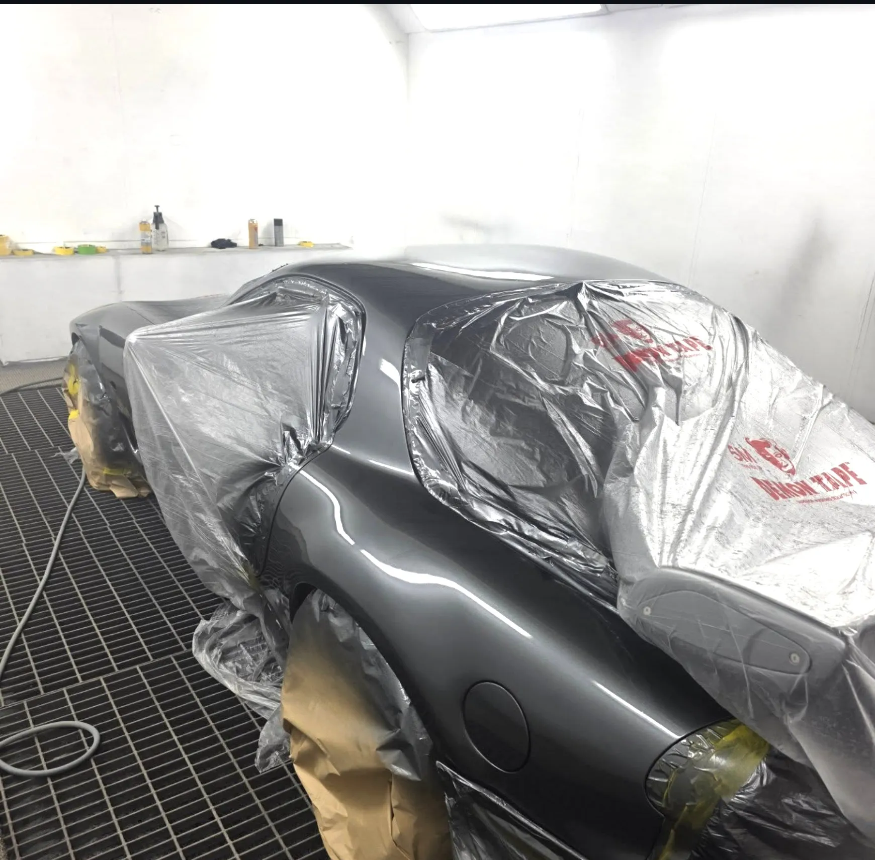 Grey Mazda RX-7 wrapped in masking film inside the spray booth, with the rear quarter panel left exposed.