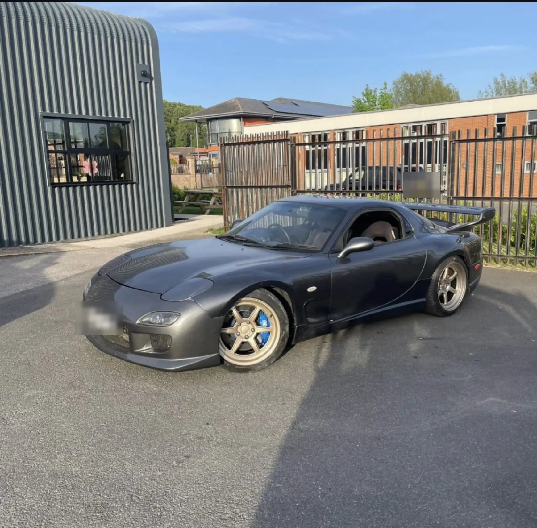 Dark grey Mazda RX-7 with a rear spoiler and bronze split-rim wheels, parked on tarmac outside a corrugated steel unit in evening sunlight.