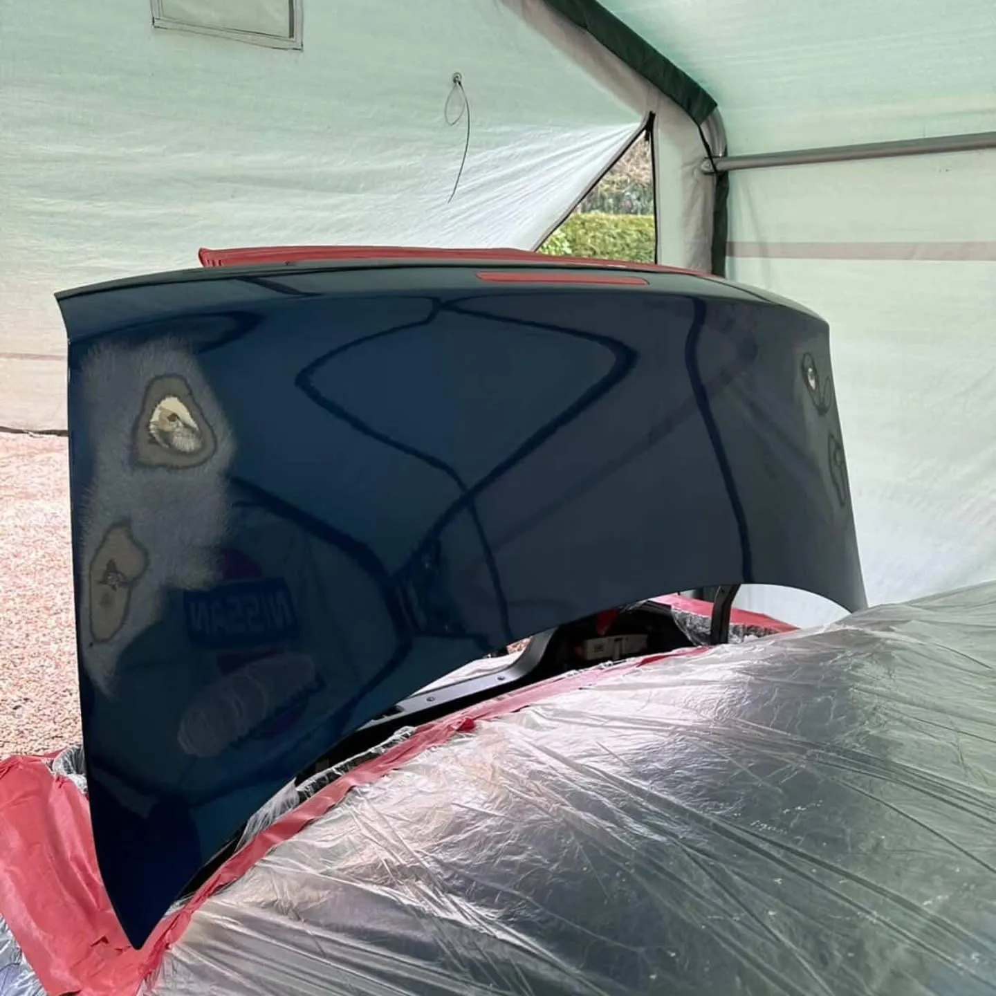 Freshly painted dark blue Nissan S14 boot lid propped open in a spray shelter, the new paint glossy enough to mirror its surroundings, with the car masked in red paper below.
