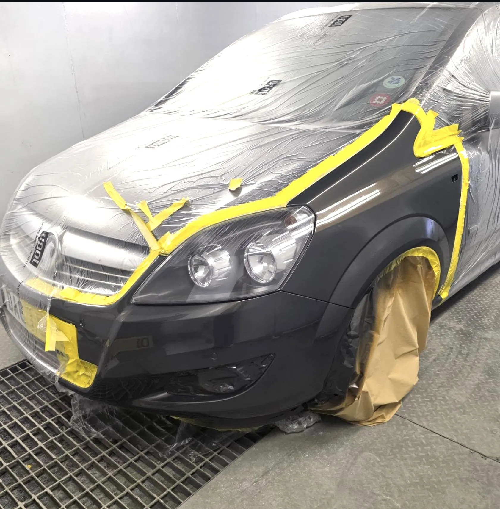Dark grey Vauxhall Zafira masked with yellow tape around the front wing and bumper corner, ready to be sprayed.